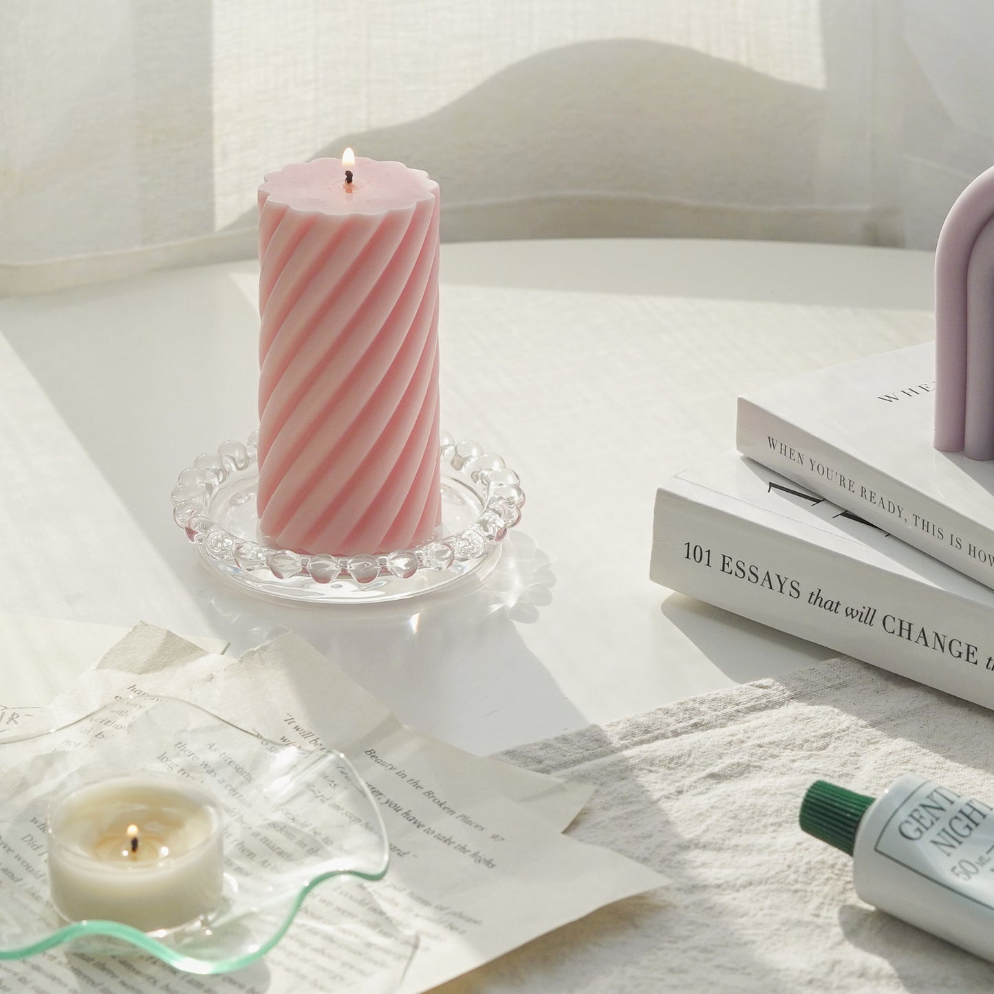 a lit blush pink sipral pillar soy candle on a clear mini beaded tray, a lit tealight candle in a ruffle dish, book pages, hand lotion, and lavender rainbow shape pillar candle on white books
