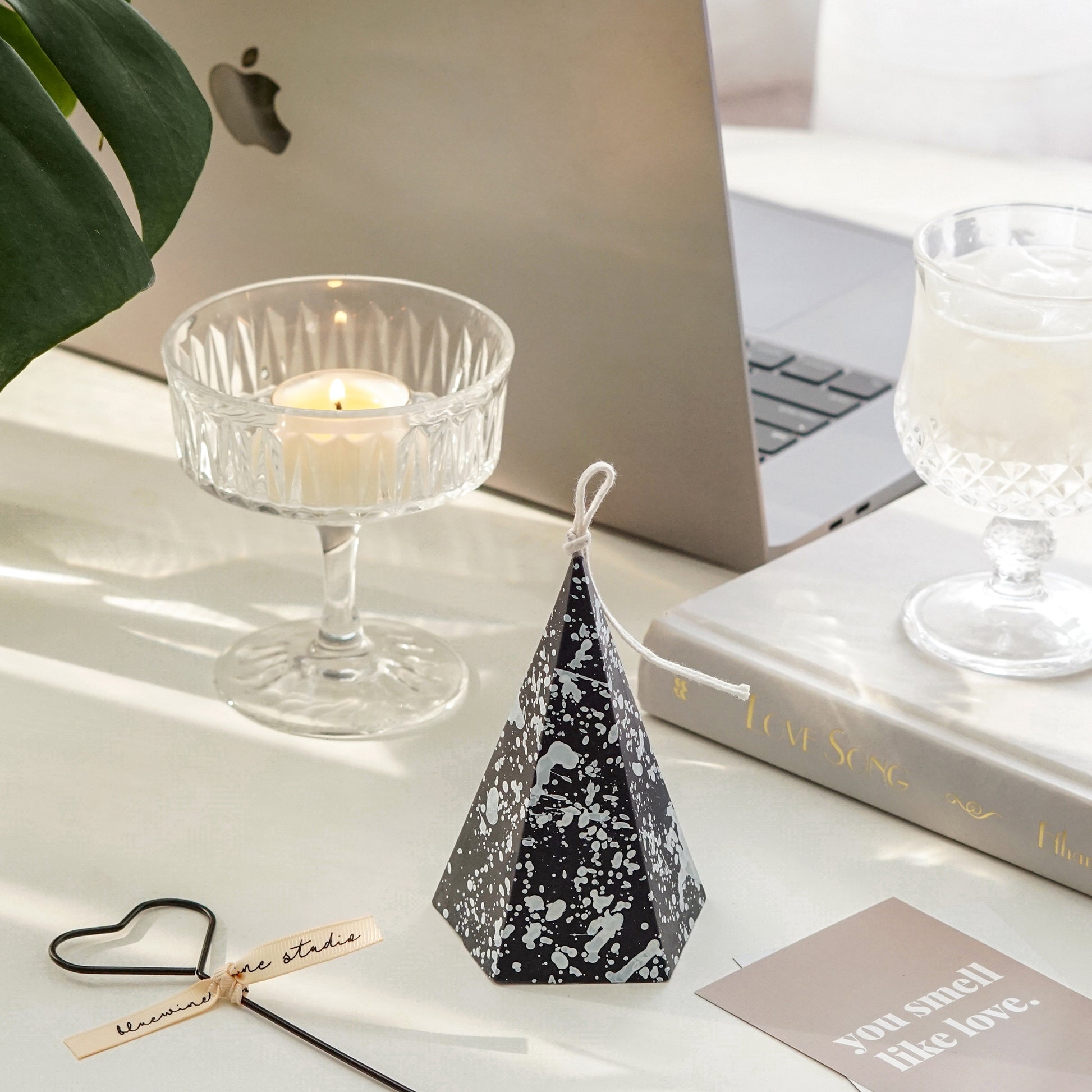 black pentagonal pyramid shape soy pillar candle with white splatter pattern, black heart shape wick dipper, you smell like love beige postcard, a lit tealight candle in a coupe glass, space gray macbook pro, a monstera leaf, mini wine glass filled with ume, and a book called love song