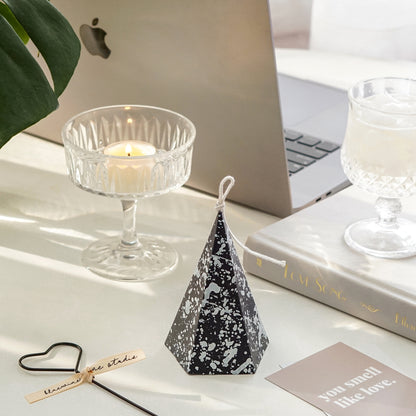 black pentagonal pyramid shape soy pillar candle with white splatter pattern, black heart shape wick dipper, you smell like love beige postcard, a lit tealight candle in a coupe glass, space gray macbook pro, a monstera leaf, mini wine glass filled with ume, and a book called love song