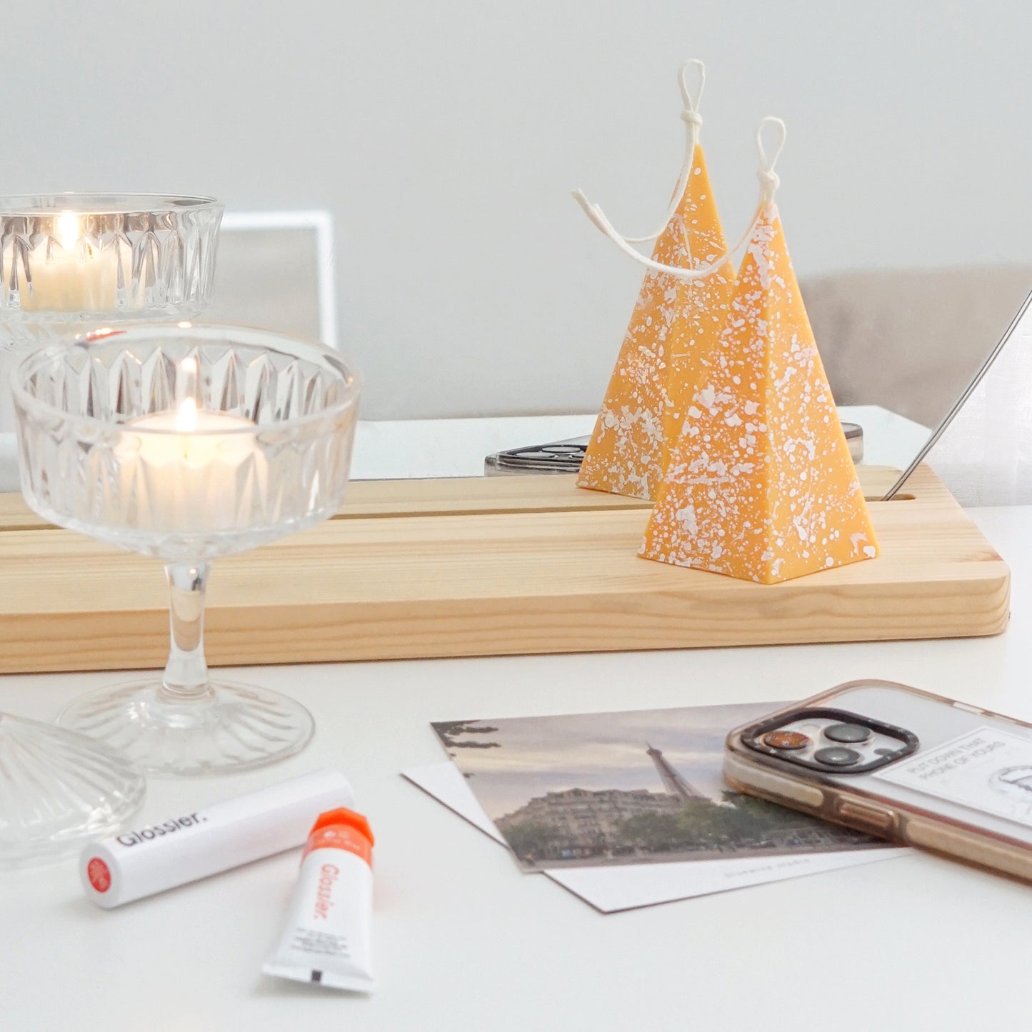 a lit pentagonal pyramid shape soy pillar candle with white splatter pattern on wavy mirror, a lit tealight candle in a coupe glass, glossier lip gloss, eiffel tower postard, and iphone 13 pro