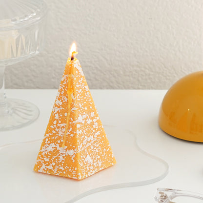 a lit yellow pentagonal pyramid shape soy pillar candle with white splatter pattern on clear wavy acrylic coaster
