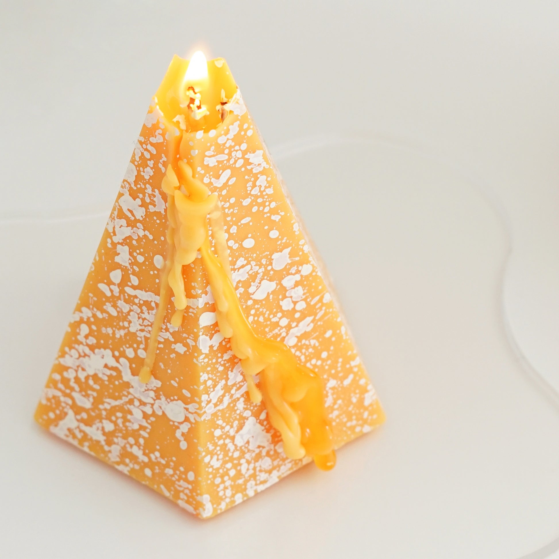 a lit yellow pentagonal pyramid shape soy pillar candle with white splatter pattern on clear wavy acrylic coaster
