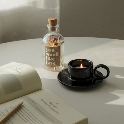 a lit tealight candle in a black teacup candle holder, a match bottle with a brown sticker inscribed with "you smell like love", and open journal with a pencil on the white round table