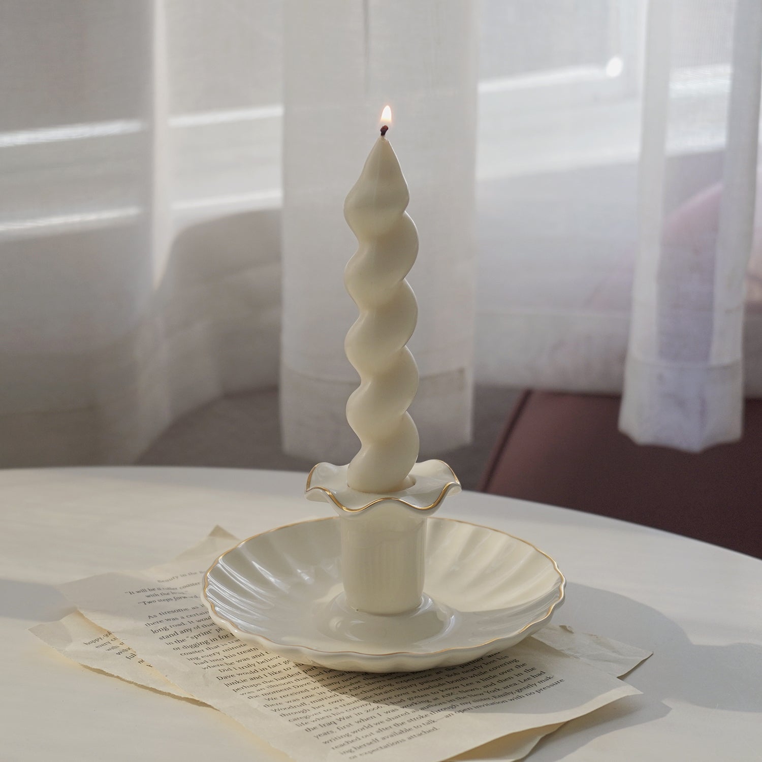 a lit white wavy spiral taper soy pillar candle in a white gold rim ruffle vintage candle holder on book pages