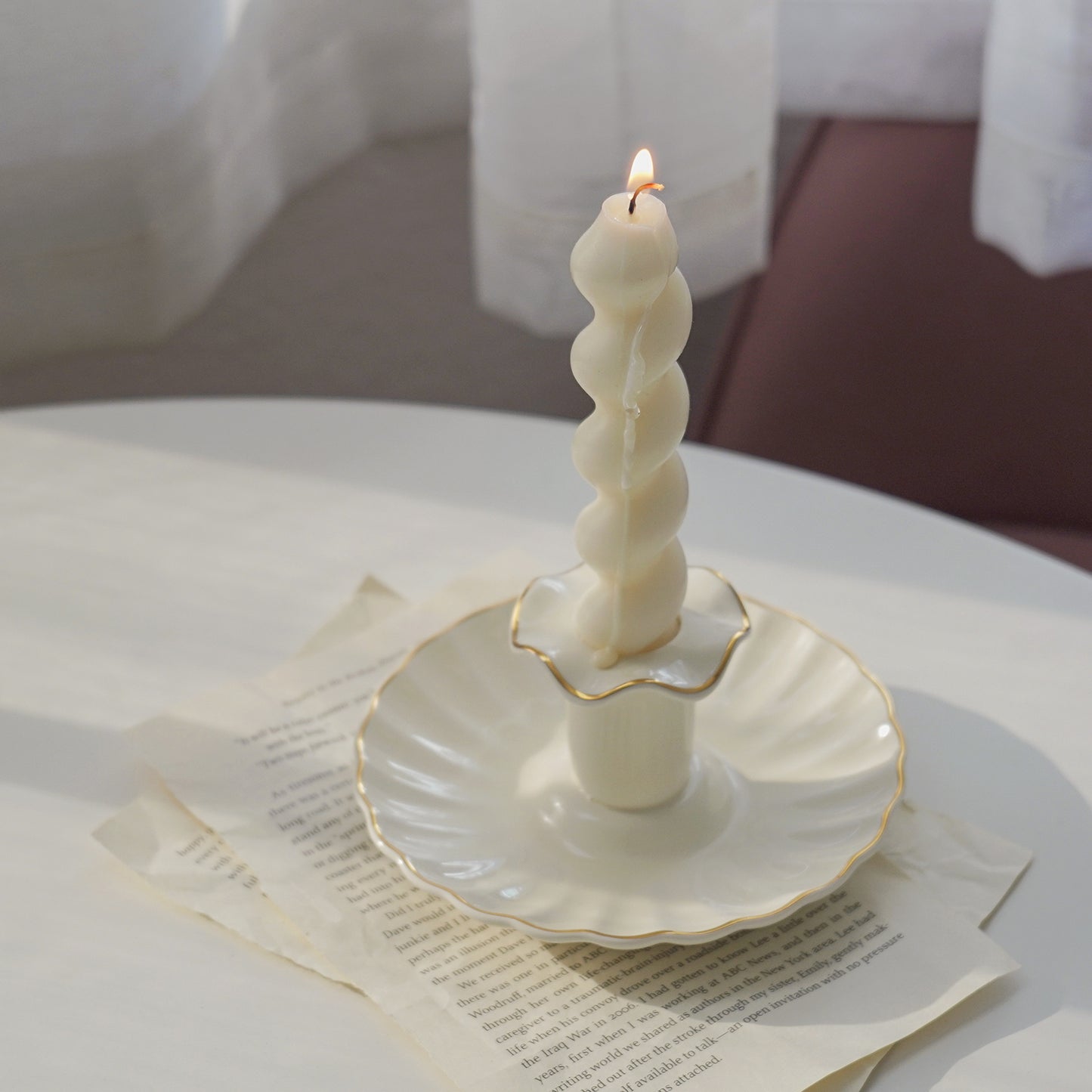 a lit white wavy spiral taper soy pillar candle in a white gold rim ruffle vintage candle holder on book pages