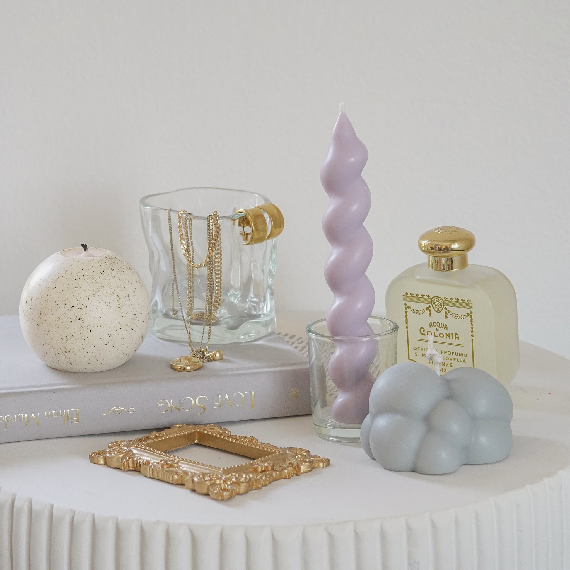 a lavender wavy spiral taper soy pillar candle in a glass, gray cloud soy candle, mini gold french picture frame, santa maria novella perfume, a dotted pattern white round sphere soy pillar candle, and jewelry hanging on wavy glass