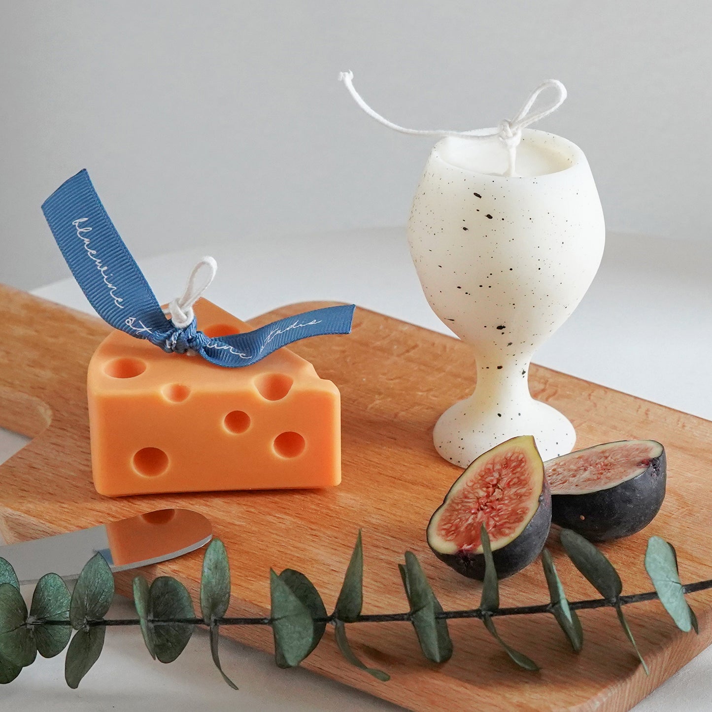 a yellow cheese soy pillar candle with blue bluewine studio ribbon, fig slices, eucalyptus leaves, cheese knife, and a dotted pattern white soy pillar wine candle on ikea mini wood board