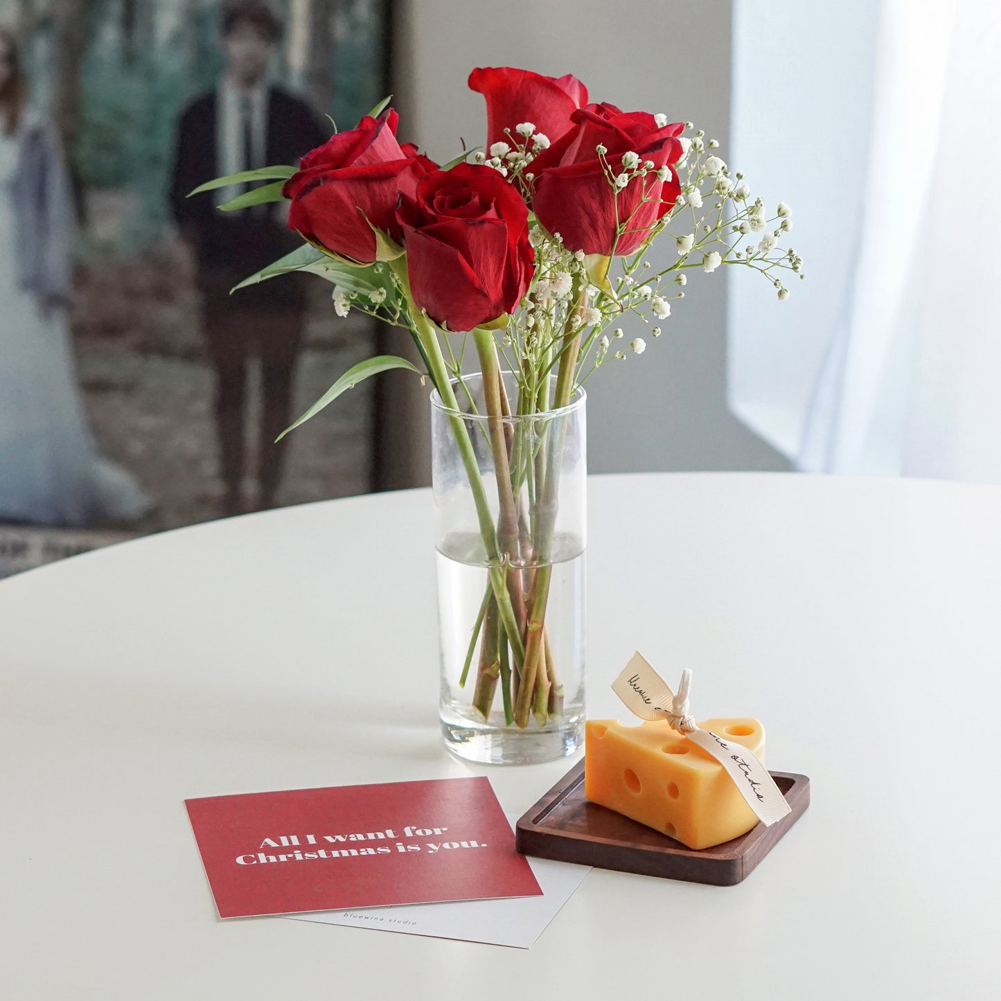 a slice of yellow cheddar cheese shape soy pillar candle on a wood square coaster, red all i want for christmas is you postcard, red roses and gypsophila in a clear cylinder vase on white round table