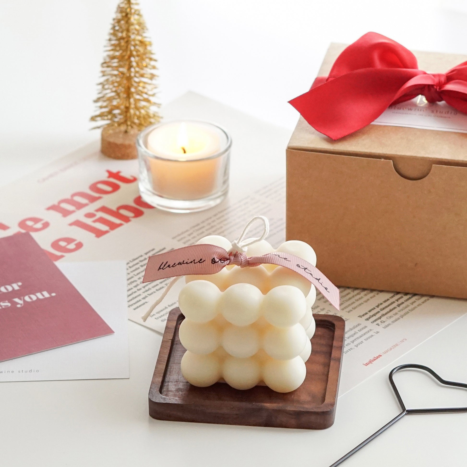 a white square cube bubble soy pillar candle on a dark brown square wood coaster, black heart shape wick dipper, burgundy postcard, magazine paper, a lit tealight candle in a glass, gold mini christmas tree decor, and kraft gift box with red ribbon