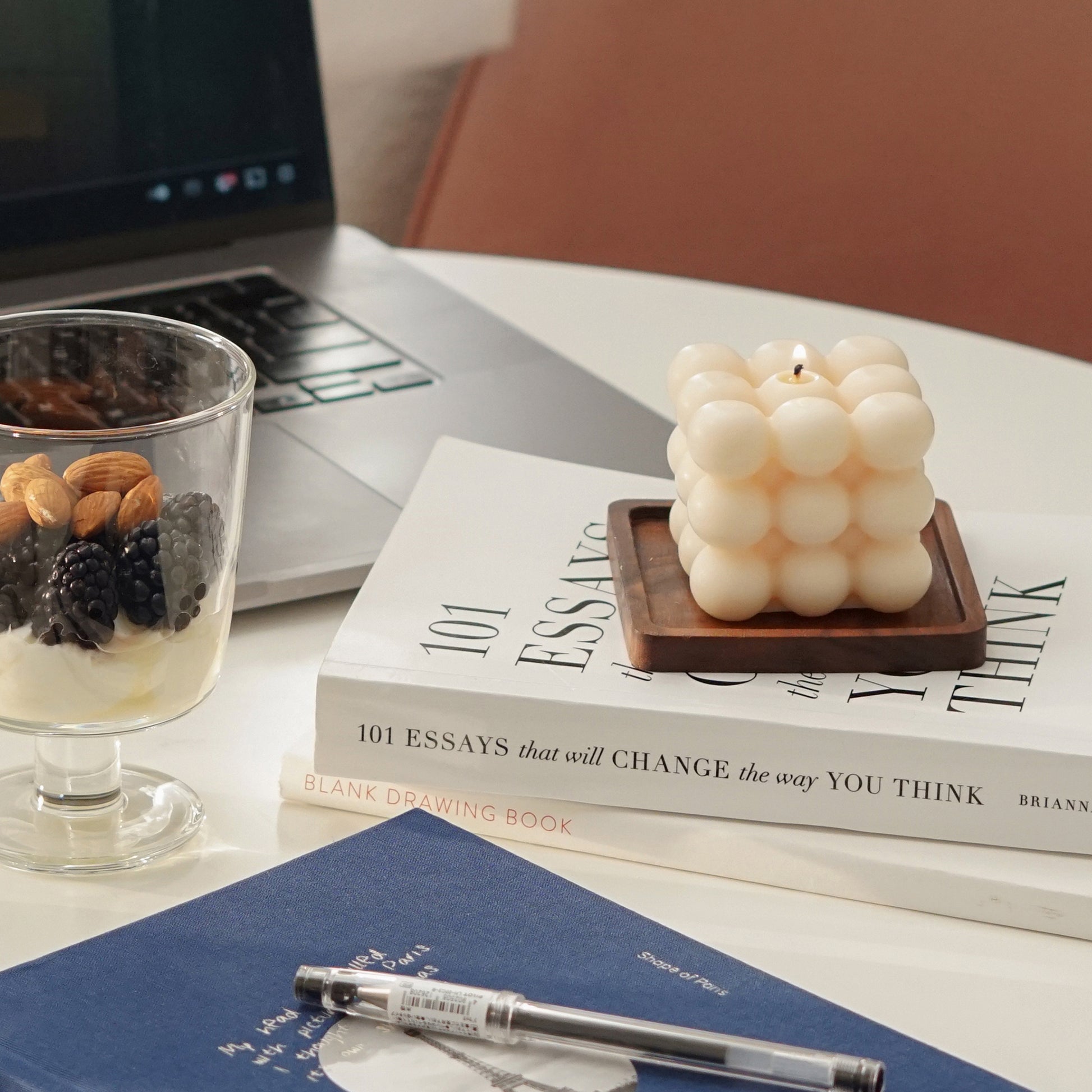 space gray macbook pro playing youtube playlist, yogurt in a tumbler glass, airpods, blue planner, a pen, a lit square cube bubble soy pillar candle on a wood square coaster placed on books