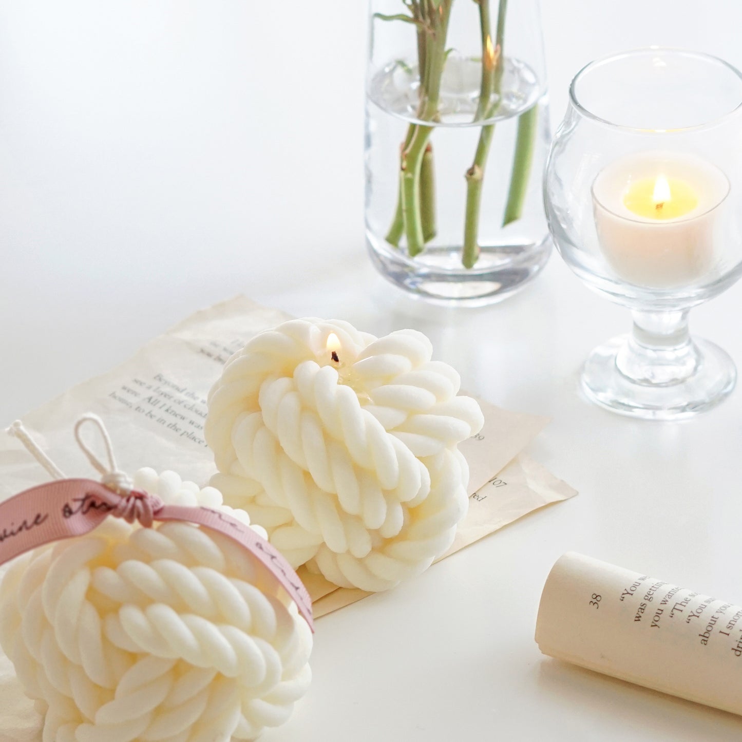 a lit yarn ball shape white soy pillar candle, another yarn candle with pink bluewine studio ribbon, rolled book pages, and a lit tealight candle in a mini glass