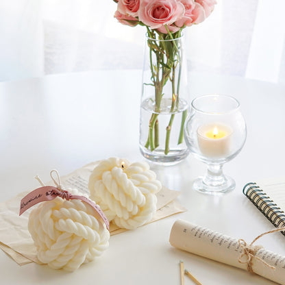 a lit yarn ball shape white soy pillar candle, another yarn candle with pink bluewine studio ribbon, rolled book pages, a note, a pink pen, pink roses in a clear vase, and a lit tealight candle in a mini glass
