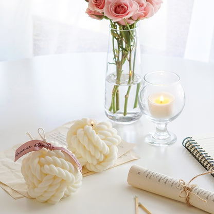 a lit yarn ball shape white soy pillar candle, another yarn candle with pink bluewine studio ribbon, rolled book pages, a note, a pink pen, pink roses in a clear vase, and a lit tealight candle in a mini glass