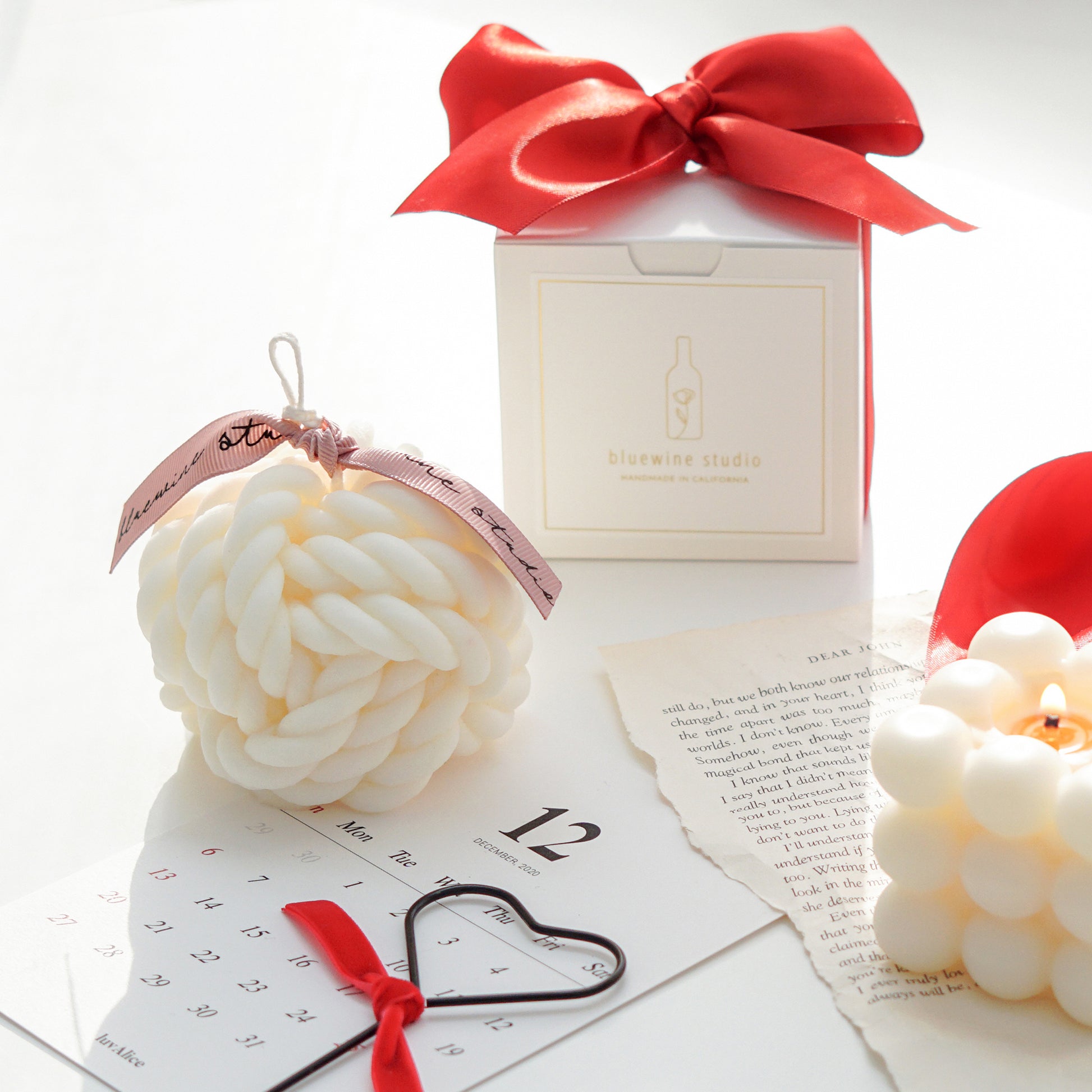 a yarn ball shape white soy pillar candle with pink bluewine studio ribbon, black heart shape wick dipper, december calendar postcard, a lit square bubble cube soy candle on a book page, and a white square bluewine studio box with red ribbon