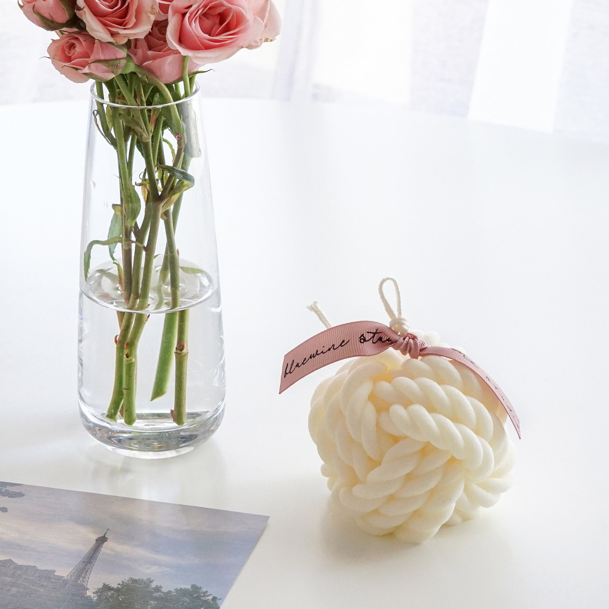 a yarn ball shape white soy pillar candle with pink bluewine studio ribbon, eiffel tower postcard, and pink roses in a clear vase