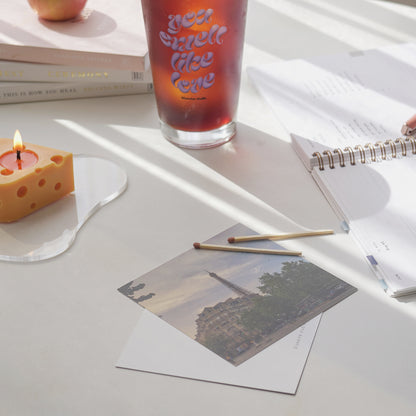 iced coffee in a 16 oz you smell like love glass, a lit cheese candle on a clear irregular shaped acrylic coaster, planner, eiffel tower postcards, and matches on the table