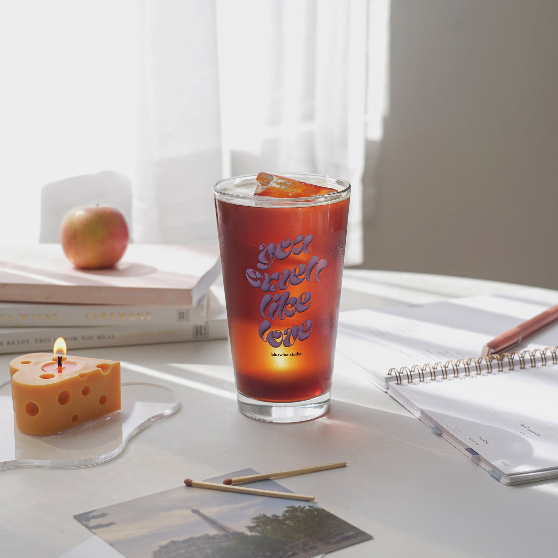 iced coffee in a 16 oz you smell like love glass, a lit cheese candle on a clear irregular shaped acrylic coaster, planner, apple, and books on the table